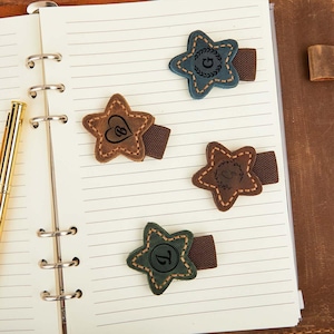 May include: Four star-shaped leather bookmarks in shades of brown, green, and blue, each with a unique stitched design and elastic band. The bookmarks feature initials and symbols, perfect for journals and notebooks.