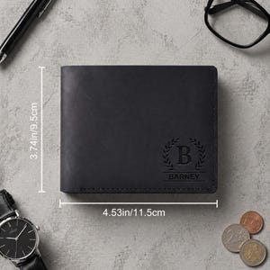Pu&ograve; includere: Un portafoglio in pelle nera, di 11,5 cm per 9,5 cm, con il nome "Barney" impresso sotto una corona d'alloro e la lettera "B". Sono visibili anche un orologio, una penna, occhiali e monete.