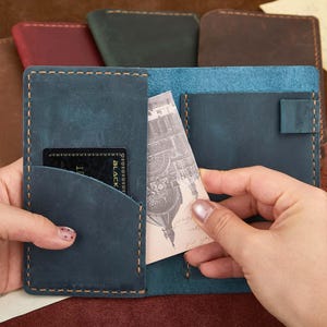 May include: A teal leather wallet with a card and a printed card inside. The wallet has a stitched border and a pocket for cards. Other leather wallets in various colors are visible in the background.