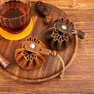 May include: Two small, brown leather pouches with drawstring closures and brass button accents sit on a wooden tray. A glass of amber liquid and a yellow coaster are in the background. A cigar is visible on the left.