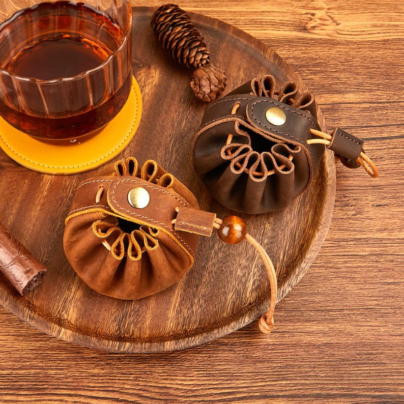 May include: Two small, brown leather pouches with drawstring closures and brass button accents sit on a wooden tray. A glass of amber liquid and a yellow coaster are in the background. A cigar is visible on the left.