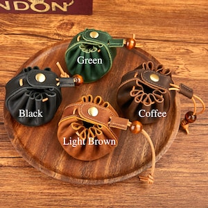 May include: Four small leather pouches in black, green, light brown, and coffee colors, each with a drawstring closure and a wooden bead accent. The pouches are displayed on a wooden tray, with the color names printed on each pouch.