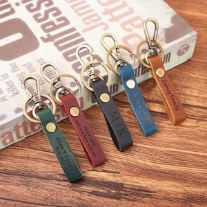 Puede incluir: Varios llaveros de cuero personalizados en colores verde, rojo, negro, azul y marrón. Cada llavero tiene un cierre metálico y una correa de cuero con texto grabado, incluyendo nombres, fechas y frases. Los llaveros están sobre una superficie de madera.
