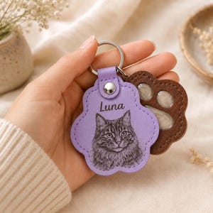 May include: A lavender paw-shaped keychain featuring a detailed black and white illustration of a cat and the name Luna printed above it. The keychain includes a silver metal ring and a secondary brown paw-shaped piece with a clear window insert.