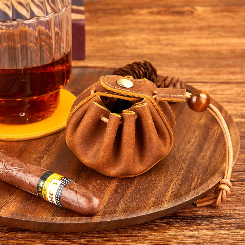May include: A brown leather pouch with a drawstring closure and a wooden bead. The pouch is sitting on a wooden tray with a cigar and a glass of amber liquid. The pouch has a snap closure.