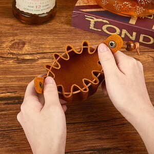 May include: A brown leather coaster with a unique wavy edge design. The coaster is held open, showcasing its interior. The leather has a natural, textured appearance. A small metal snap secures the coaster's handle.
