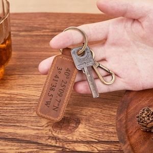 Puede incluir: Un llavero de cuero marrón con coordenadas grabadas y un llavero de latón. El llavero rectangular tiene bordes cosidos y las coordenadas "40°25'09.7"N 3°41'38.5"W" son visibles. Una llave y un mosquetón están adjuntos.