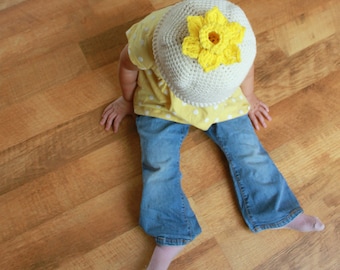 May include: A child wearing a yellow and white crocheted hat shaped like a daffodil flower. The hat has a yellow flower on top and is sitting on the child's head. The child is wearing blue jeans and a yellow shirt with white polka dots.