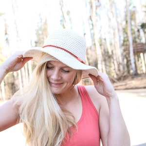 May include: A woman wearing a white crochet sun hat with a brown leather band and a coral tank top.