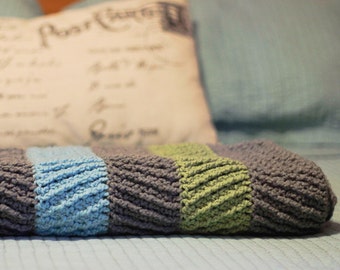 May include: A striped crocheted throw blanket in shades of gray, blue, and green, folded on a blue bedspread.