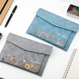Peut inclure: Deux pochettes en similicuir, une grise et une bleue, avec des motifs floraux et le nom "Sophie" imprimé dessus. La pochette grise a un papillon. La pochette bleue est au-dessus de la grise.