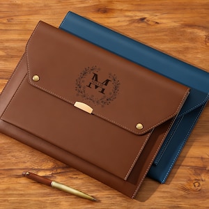 Könnte beinhalten: Eine braune Ledermappe mit goldenem Verschluss und Druckknopfverschluss, mit einem floralen Kranzdesign mit dem Buchstaben "M" und dem Namen "Millie". Eine blaue Mappe ist hinter der braunen sichtbar. Ein Stift liegt auf der Holzoberfläche.