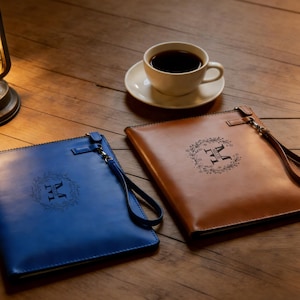 May include: Two leather clutch bags, one blue and one brown, with a floral wreath and the letter "M" engraved on the front. Each bag has a wrist strap. A cup of coffee on a saucer sits on a wooden surface.