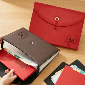 Peut inclure: Plusieurs organiseurs de documents rouges, marron et noirs. L'organiseur marron est ouvert, révélant des séparateurs étiquetés et des dossiers rouges. Un organiseur rouge avec une fermeture à rabat et un logo décoratif est également visible.