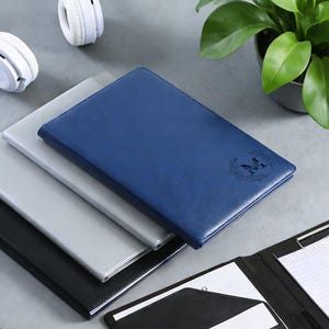 Peut inclure: Trois portfolios en cuir empilés, de couleurs bleu, gris et noir. Le portfolio bleu présente un emblème décoratif. Un casque blanc et une plante en pot sont en arrière-plan. Les portfolios sont fermés.