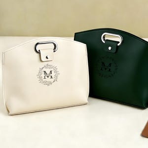 May include: Two leather tote bags, one cream and one dark green, with silver handles. Each bag features a monogram with the letter "M" inside a wreath and the name "Millie" below. The bags have a structured design.