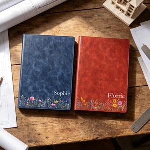 May include: Two rectangular notebooks with a textured finish, one in deep blue and one in reddish-brown. Each features a colourful wildflower print along the bottom edge and a name printed in white: Sophie on the blue notebook and Florrie on the red.