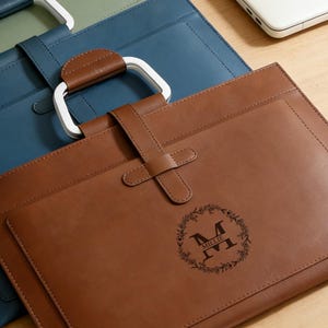 May include: Brown leather laptop case with a white handle and a strap closure. The case features a monogram with the letter "M" and the name "Millie" inside a floral wreath. A blue leather case is visible in the background.