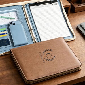 May include: A brown leather-effect zipped portfolio featuring the name Catherine and a laurel wreath logo embossed on the cover. The interior is light blue, containing a metal ring binder, a memo pad, a smartphone pocket, and various card slots.