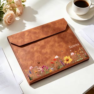 May include: A brown leather-look document folder featuring a flap closure and a printed design of colourful wildflowers, green stems, and a small butterfly. The name Serena is printed in white script on the front right side of the folder.