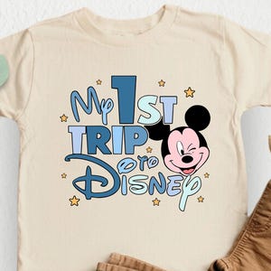 K&ouml;nnte beinhalten: Ein beiges T-Shirt mit einer blauen und wei&szlig;en Grafik, auf der "My 1st Trip to Disney" steht, sowie einem Cartoon-Bild von Mickey Mouse. Das Shirt liegt auf einem wei&szlig;en Hintergrund mit einer braunen Hose und einer gr&uuml;nen Pflanze.