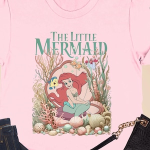 May include: Pink t-shirt featuring "The Little Mermaid" graphic, paired with dark blue jeans, a black purse, and black flats. The shirt has rolled-up sleeves and the graphic shows Ariel in a seashell.
