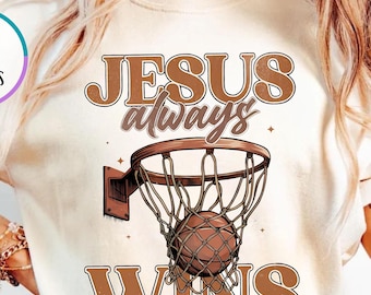 May include: Ivory-colored t-shirt featuring a basketball hoop with a ball in the net. The text "JESUS always WINS" is printed in brown. The shirt has short sleeves and a round neckline.