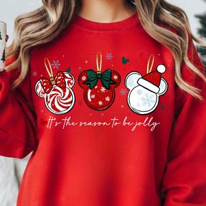 May include: Red sweatshirt with three Mickey Mouse-shaped Christmas ornament designs. The ornaments feature a candy cane, a red bow, and a Santa hat. The text "It's the season to be jolly" is printed below the ornaments. A white mug with Christmas trees is held in the hand.