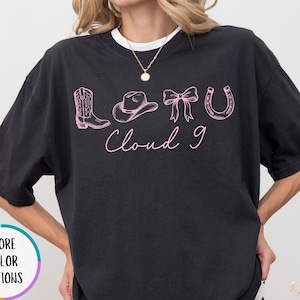 May include: Black t-shirt with a white collar featuring a pink graphic design of a cowboy boot, hat, bow, and horseshoe, with the text "Cloud 9". The shirt is a relaxed fit.