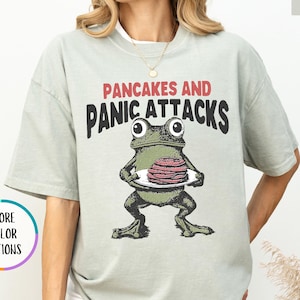 May include: A light green t-shirt with the text "PANCAKES AND PANIC ATTACKS" above a cartoon frog holding a plate of pancakes. The shirt has short sleeves and a relaxed fit.