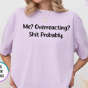 May include: An orchid-colored short-sleeve t-shirt featuring the black text "Me? Overreacting? Shit Probably." printed on the chest. The shirt has a crew neckline and a relaxed fit, paired with black shorts. A small circular graphic indicates more color options.
