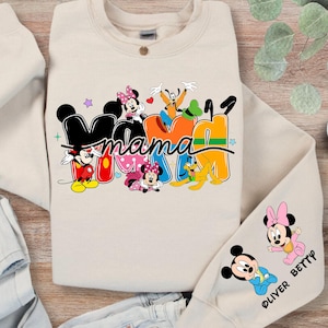 K&ouml;nnte beinhalten: Ein sandfarbenes Sweatshirt mit dem Wort "Mama" in einem farbenfrohen Disney-Design. Das Design umfasst Micky und Minnie Maus, Goofy und Pluto. Der &Auml;rmel hat zus&auml;tzliche Minnie- und Micky-Maus-Grafiken mit dem Text "OLIVER BETTY".