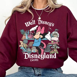 May include: Maroon sweatshirt featuring a graphic of Minnie Mouse and the text "Walt Disney's Disneyland Est.1955". The design includes various Disney park icons. The sweatshirt is a solid maroon color.