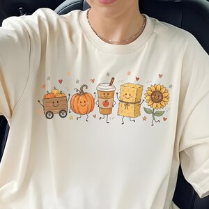 May include: A cream-colored t-shirt featuring a graphic print of five animated autumn-themed characters in a row: a wagon with pumpkins, a single pumpkin, a coffee cup, a hay bale, and a sunflower, all accented by small hearts and stars.