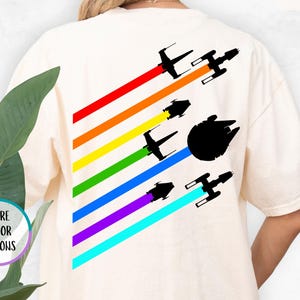 K&ouml;nnte beinhalten: Ein wei&szlig;es T-Shirt mit einem Regenbogenmuster und Silhouetten von Star Wars Raumschiffen. Der Text "IVORY" befindet sich in der oberen rechten Ecke des Bildes.