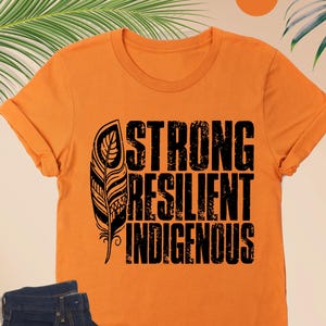 May include: Orange t-shirt with the text "STRONG RESILIENT INDIGENOUS" and a feather graphic. The shirt is paired with dark blue jeans, eyeglasses, and cream-colored moccasins.