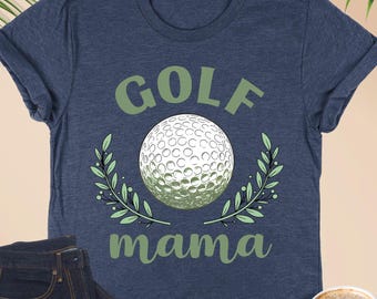 May include: Heather navy blue t-shirt with the text "GOLF mama" in green, featuring a golf ball and laurel leaves. A pair of dark blue jeans, a cup of coffee, and eyeglasses are also in the image.
