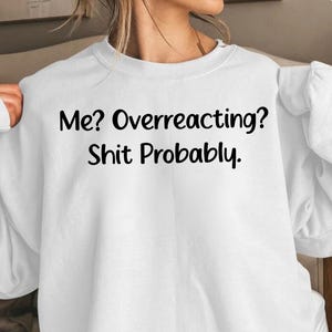 May include: A white long-sleeve sweatshirt featuring the black printed text Me? Overreacting? Shit Probably. The garment has a crew neckline, long sleeves with ribbed cuffs, and a relaxed fit, providing a simple and clean look for casual wear.