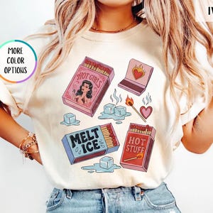 May include: Ivory-colored t-shirt featuring a graphic design with matchboxes and ice cubes. The matchboxes have text that reads "Hot Girls", "Melt Ice", and "Hot Stuff". The shirt also has the text "More Color Options".