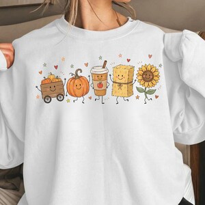 May include: A white long-sleeve sweatshirt featuring a graphic print of five cartoon characters: a wooden wagon with pumpkins, an orange pumpkin, a coffee cup, a hay bale, and a sunflower, all accented with small red hearts and stars.