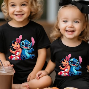 K&ouml;nnte beinhalten: Zwei Kinder tragen schwarze T-Shirts mit einer bunten Grafik von Lilo und Stitch. Die Grafik zeigt Lilo in einem roten Kleid und Stitch in Blau. Die Kinder l&auml;cheln. Die T-Shirts sind kurz&auml;rmlig.