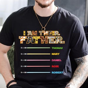 Puede incluir: Camiseta negra con el texto "I AM THEIR FATHER" en amarillo y blanco, con nombres y dise&ntilde;os de sables de luz debajo. Se enumeran los nombres Thomas, Mary, Daniel, Rose y Robert. La camiseta es de manga corta.