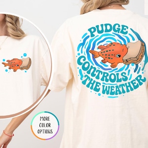 Puede incluir: Camiseta de color marfil con un dise&ntilde;o de pez de dibujos animados. La parte delantera de la camiseta tiene un peque&ntilde;o gr&aacute;fico de pez, mientras que la parte trasera muestra un dise&ntilde;o m&aacute;s grande con el texto "Pudge Controls the Weather" en azul. La camiseta es de corte holgado.