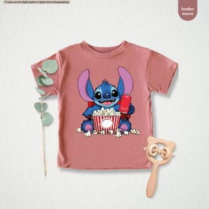 May include: Heather mauve t-shirt featuring a cartoon Stitch character holding a red soda can and a popcorn bucket. The blue Stitch is smiling, surrounded by popcorn. The shirt is a short-sleeved crew neck.