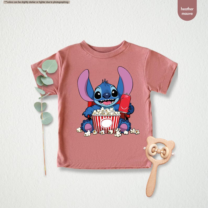 May include: Heather mauve t-shirt featuring a cartoon Stitch character holding a red soda can and a popcorn bucket. The blue Stitch is smiling, surrounded by popcorn. The shirt is a short-sleeved crew neck.