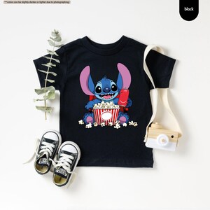 May include: Black t-shirt featuring a cartoon character, holding a red soda can and a popcorn bucket. The character is blue with large ears. White popcorn kernels are scattered around the bucket. Black text in the upper right corner says "black".