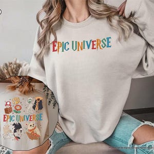 May include: A sand-colored sweatshirt with the text "EPIC UNIVERSE" in colorful, stylized lettering. The design includes various cartoon characters. The sweatshirt is a casual, comfortable garment.