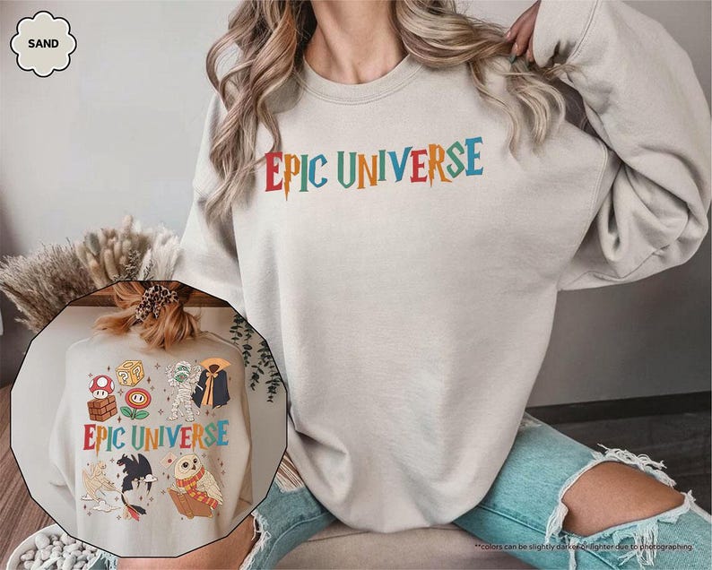 May include: A sand-colored sweatshirt with the text "EPIC UNIVERSE" in colorful, stylized lettering. The design includes various cartoon characters. The sweatshirt is a casual, comfortable garment.