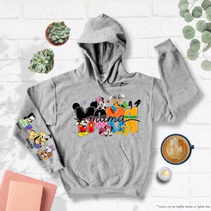 K&ouml;nnte beinhalten: Ein grauer Kapuzenpullover mit dem Wort "Mama" in einem farbenfrohen Disney-Design. Der linke &Auml;rmel zeigt Cartoon-Figuren und Namen. Der Pullover hat eine Kapuze und eine Vordertasche. Er ist aus weichem Material.