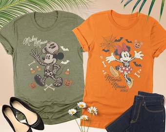 May include: A heather olive t-shirt featuring a skeleton Mickey Mouse graphic with bats and candy corn, and an orange t-shirt featuring a skeleton Minnie Mouse graphic with a spiderweb, bats, and pumpkins, paired with dark blue denim shorts.